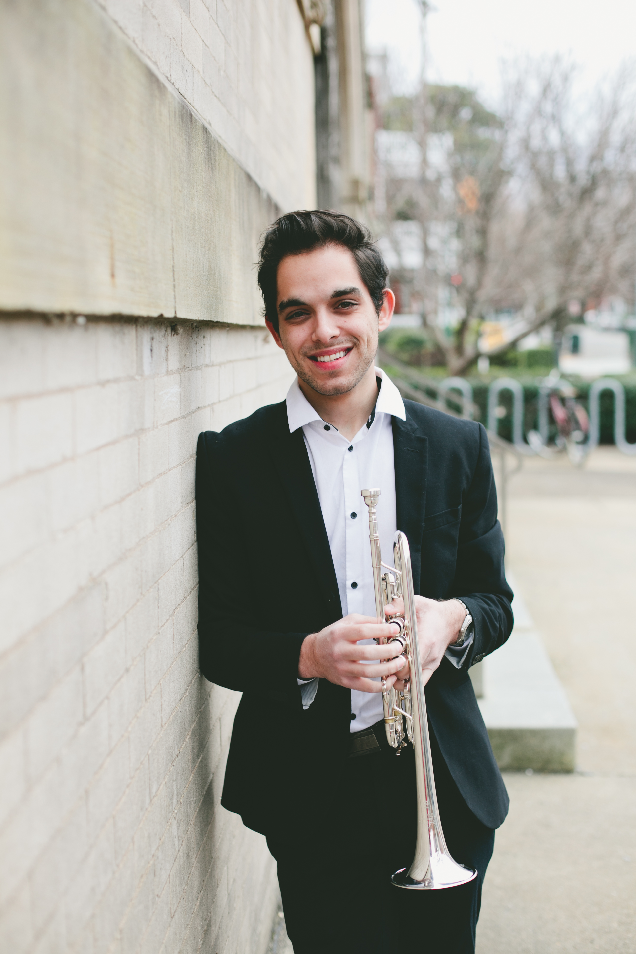 Adjunct Instructor Haidar leaning against a brick wall holding a trumpet Adjunct Instructor Haidar leaning against a brick wall holding a trumpet